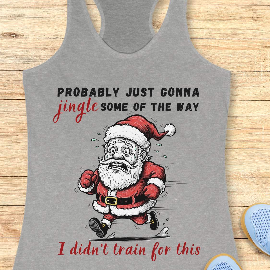 A grey racerback tank top laid flat on a light wood background, featuring a humorous graphic of a running Santa Claus with the text 'PROBABLY JUST GONNA jingle SOME OF THE WAY I didn't train for this'.