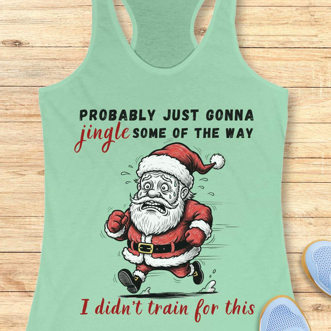 A mint green racerback tank top laid flat on a light wood background, featuring a humorous graphic of a running Santa Claus with the text 'PROBABLY JUST GONNA jingle SOME OF THE WAY I didn't train for this'.