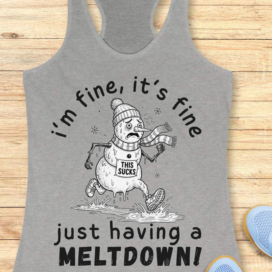 A heather gray racerback tank top featuring a black and white graphic of a melting snowman running in a puddle with the text 'i'm fine, it's fine just having a MELTDOWN!'. The tank top is displayed flat on a light wood background with blue sneakers partially visible in the corner.