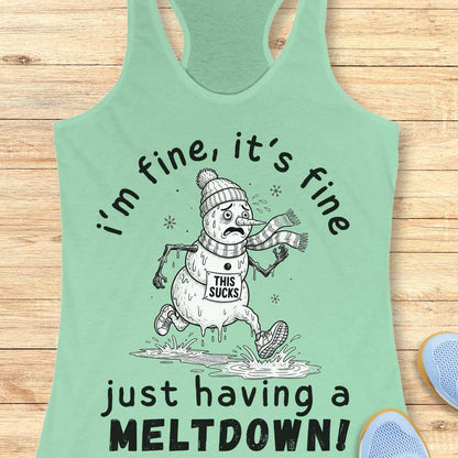 A mint green racerback tank top with a humorous snowman graphic and the text 'i'm fine, it's fine just having a MELTDOWN!' displayed on a light wood background with blue sneakers partially visible.