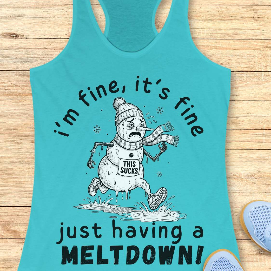 A turquoise racerback tank top laid flat on a light wood surface, featuring a cartoon snowman graphic with the text 'i'm fine, it's fine just having a MELTDOWN!'. A partial blue shoe is visible at the bottom right as a styling prop.