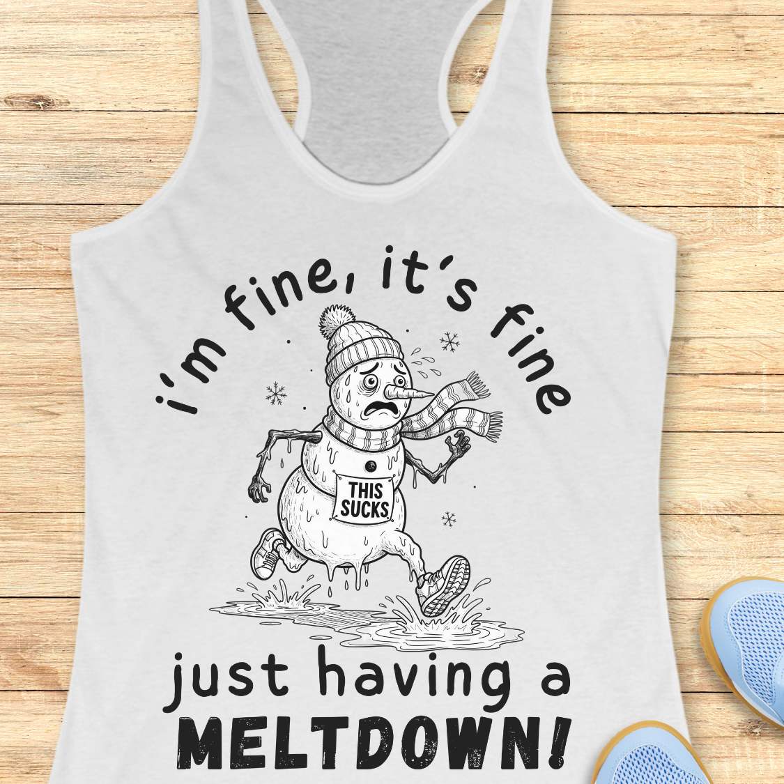 A white racerback tank top laid flat on a light wood background, featuring a black-and-white winter snowman graphic with the text 'i'm fine, it's fine just having a MELTDOWN!'. A small blue shoe is partially visible at the bottom right as a styling prop.