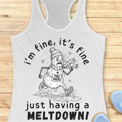 A white racerback tank top laid flat on a light wood background, featuring a black-and-white winter snowman graphic with the text 'i'm fine, it's fine just having a MELTDOWN!'. A small blue shoe is partially visible at the bottom right as a styling prop.