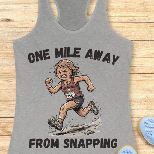Heather gray racerback tank top displayed on a light wooden surface, featuring a humorous running-themed graphic and bold black text. The design shows a determined runner wearing a race bib numbered “66” and a tank top labeled “THE GRIND,” sprinting along a rocky trail with dust and debris kicking up beneath running shoes. Large text above and below the illustration reads: “ONE MILE AWAY” and “FROM SNAPPING.” The artwork uses a cartoon-style illustration with distressed typography, capturing the relatable r