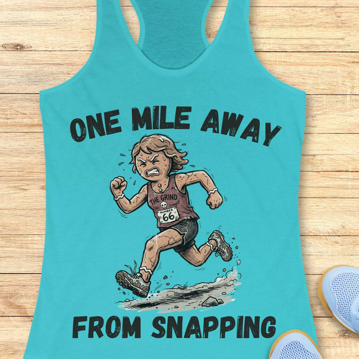 blue racerback tank top displayed on a light wooden surface, featuring a humorous running-themed graphic and bold black text. The design shows a determined runner wearing a race bib numbered “66” and a tank top labeled “THE GRIND,” sprinting along a rocky trail with dust and debris kicking up beneath running shoes. Large text above and below the illustration reads: “ONE MILE AWAY” and “FROM SNAPPING.” The artwork uses a cartoon-style illustration with distressed typography, capturing the relatable r