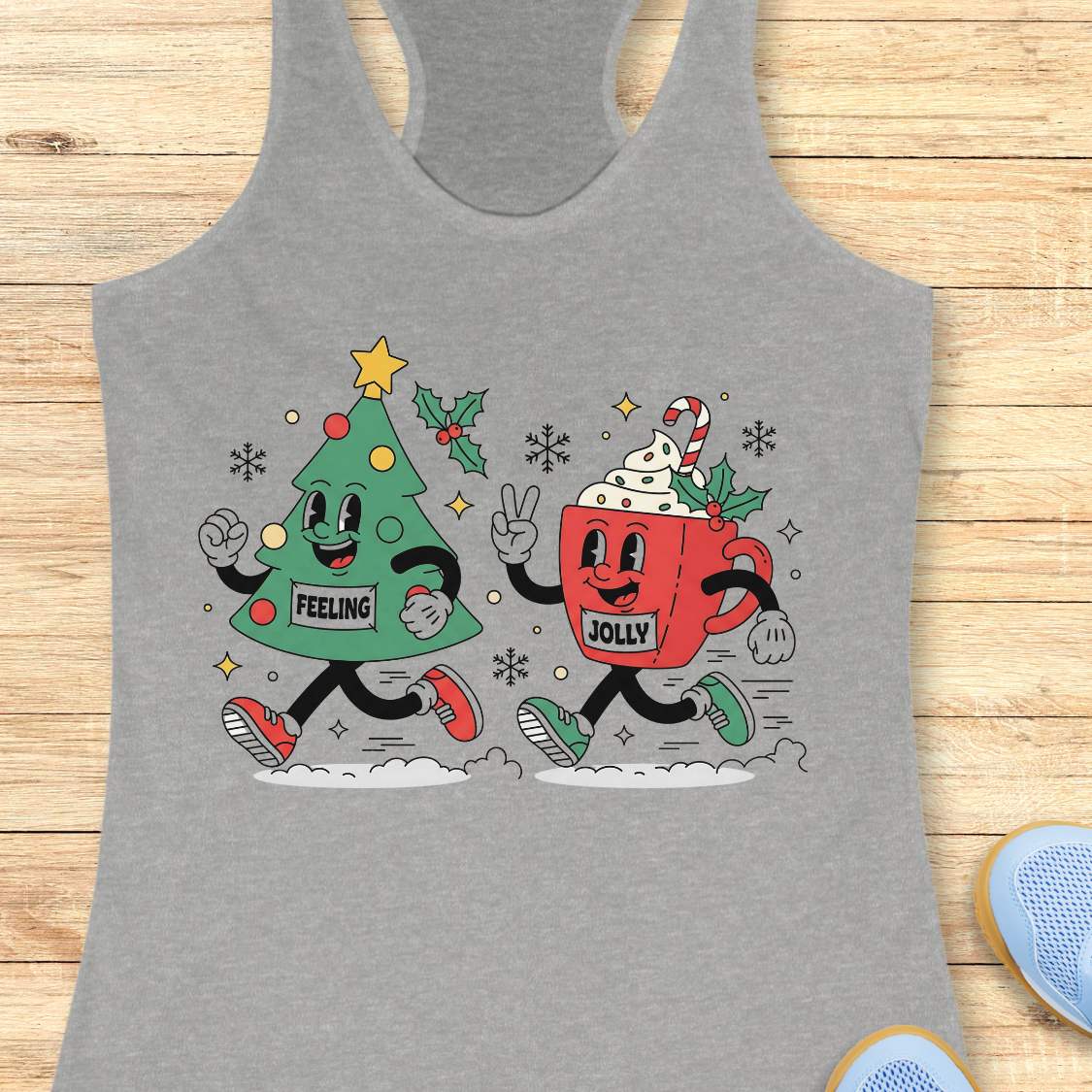 A heather gray racerback tank top laid flat on a light wood surface, featuring a colorful Christmas-themed graphic of a green Christmas tree and a red hot cocoa mug with the words 'FEELING' and 'JOLLY' on the front.