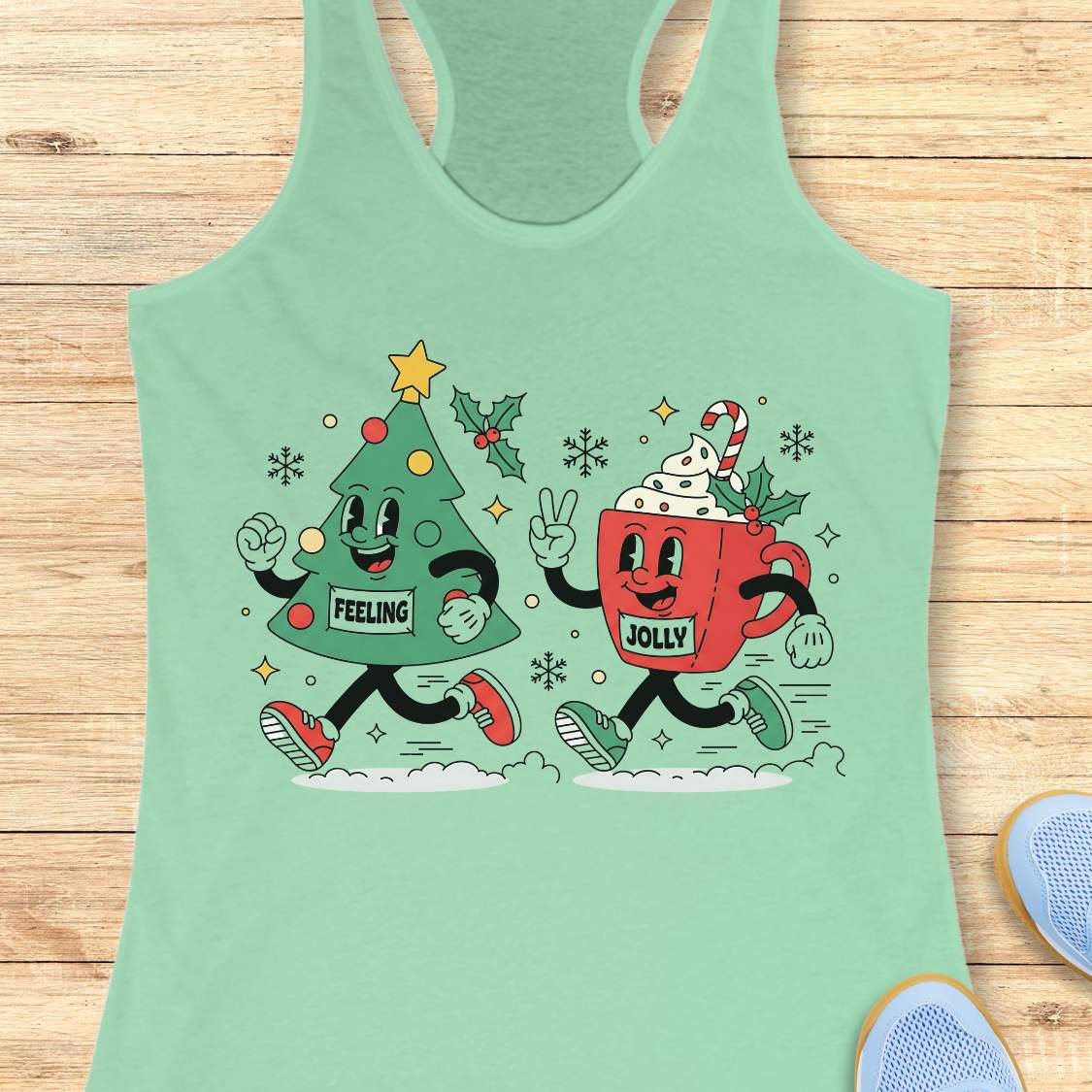 A mint green racerback tank top laid flat on a light wood background, featuring a colorful graphic of a green Christmas tree and a red hot cocoa mug running together with holiday decorations and the words 'FEELING' and 'JOLLY'.