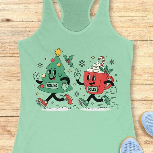 A mint green racerback tank top laid flat on a light wood background, featuring a colorful graphic of a green Christmas tree and a red hot cocoa mug running together with holiday decorations and the words 'FEELING' and 'JOLLY'.