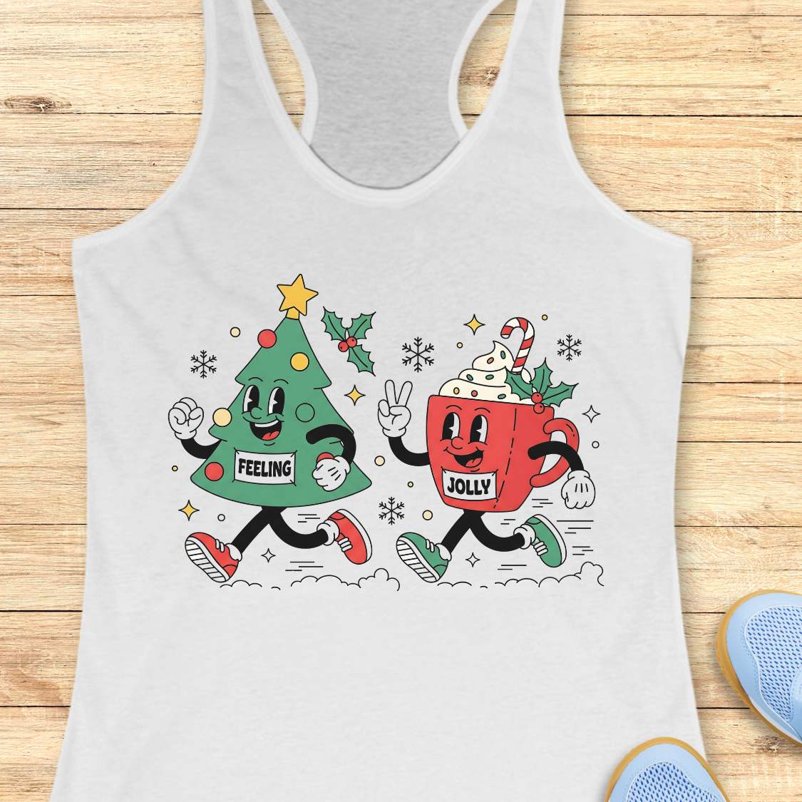 A white racerback tank top laid flat on a light wood background, featuring a colorful graphic of a green Christmas tree and a red hot cocoa mug with the words 'FEELING JOLLY' and festive illustrations.
