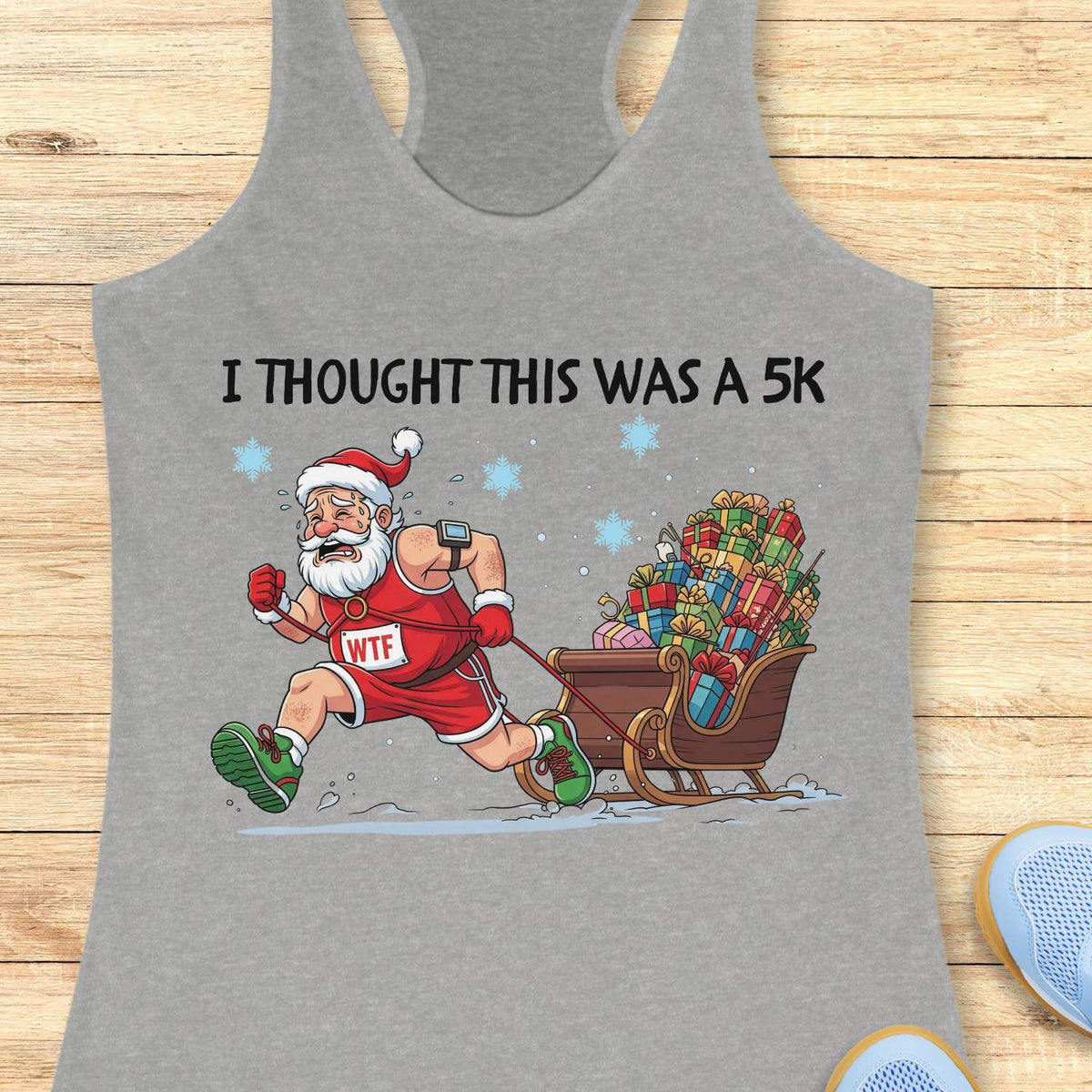 grey racerback tank top displayed on a light wooden surface, featuring a humorous Christmas running graphic. The design shows Santa Claus dressed in a red running outfit, Santa hat, gloves, and green running shoes, pulling a sleigh overflowing with colorful wrapped presents while running through the snow. Santa wears a race bib labeled “WTF,” adding a playful touch to the design. White text above the illustration reads: “I THOUGHT THIS WAS A 5K.” Blue snowflakes and snowy ground details complete the festiv