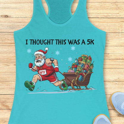 blue racerback tank top displayed on a light wooden surface, featuring a humorous Christmas running graphic. The design shows Santa Claus dressed in a red running outfit, Santa hat, gloves, and green running shoes, pulling a sleigh overflowing with colorful wrapped presents while running through the snow. Santa wears a race bib labeled “WTF,” adding a playful touch to the design. White text above the illustration reads: “I THOUGHT THIS WAS A 5K.” Blue snowflakes and snowy ground details complete the festiv